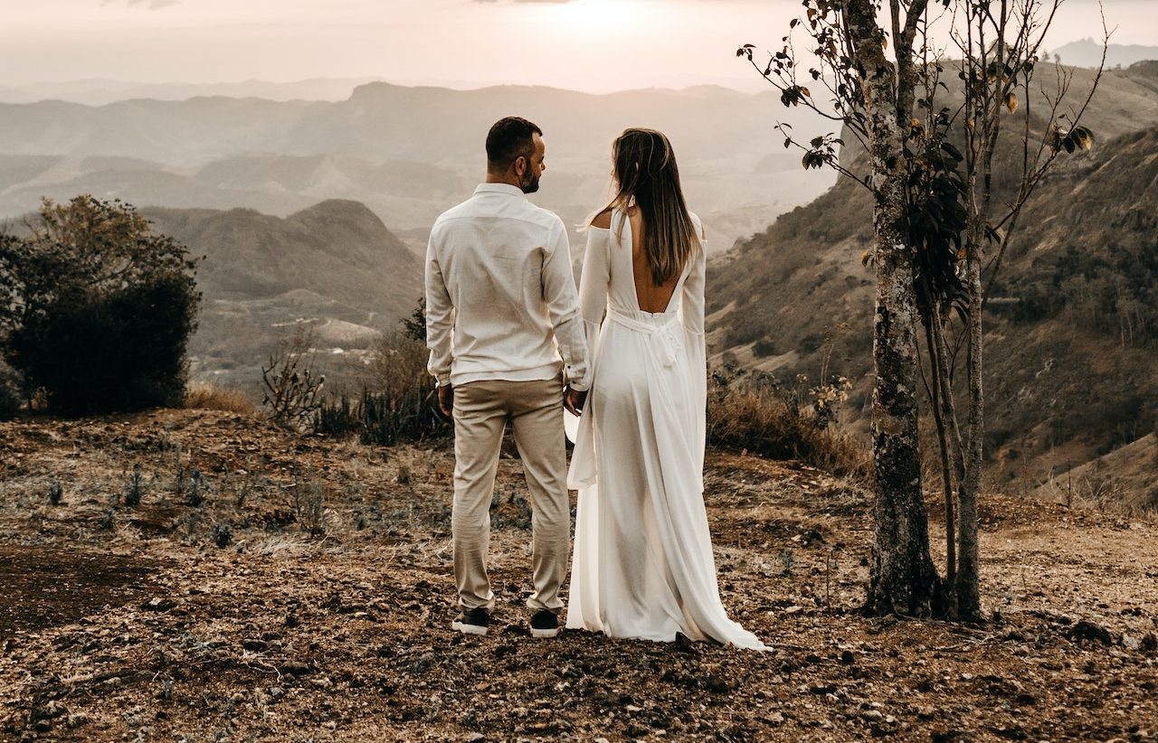A Couple In White Dress Standing In View Of The Mountain